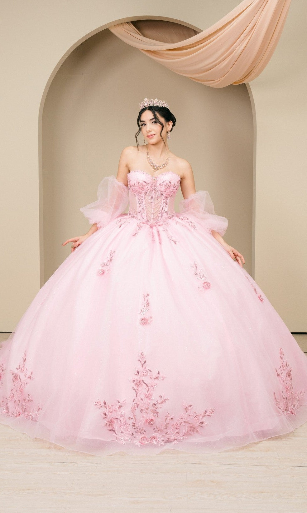 A woman wearing the Quinceanera Dress 8380J By Cinderella Couture in pink, featuring floral embroidery and tulle accents, stands before an arched backdrop with a tiara and necklace.